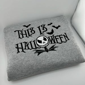 K&ouml;nnte beinhalten: Ein graues Sweatshirt mit dem schwarzen Schriftzug "THIS IS HALLOWEEN". Das Design umfasst Flederm&auml;use und eine Figur im Jack-Skellington-Stil. Das gefaltete Sweatshirt scheint aus einem weichen Stoff zu sein.
