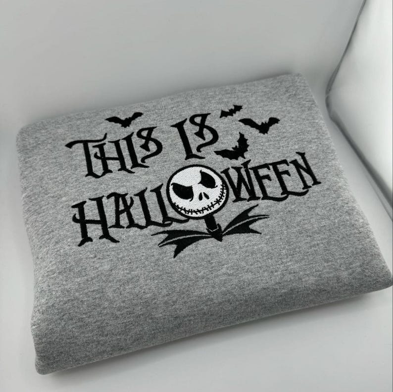 K&ouml;nnte beinhalten: Ein graues Sweatshirt mit dem schwarzen Schriftzug "THIS IS HALLOWEEN". Das Design umfasst Flederm&auml;use und eine Figur im Jack-Skellington-Stil. Das gefaltete Sweatshirt scheint aus einem weichen Stoff zu sein.