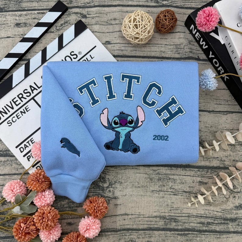 May include: A light blue sweatshirt featuring an embroidered Stitch character design. The word "STITCH" is arched across the chest in blue lettering, with the year "2002" below. A small Stitch graphic is on the left sleeve.