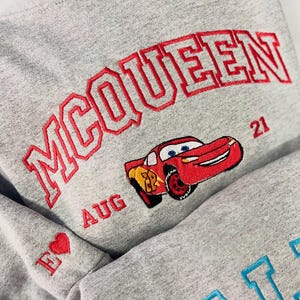 Pode incluir: Um moletom cinza com texto bordado em vermelho lendo "McQUEEN" acima de um carro de desenho animado vermelho. A manga apresenta um cora&ccedil;&atilde;o vermelho e a letra "E". A palavra "AUG" e o n&uacute;mero "21" tamb&eacute;m s&atilde;o bordados no moletom.