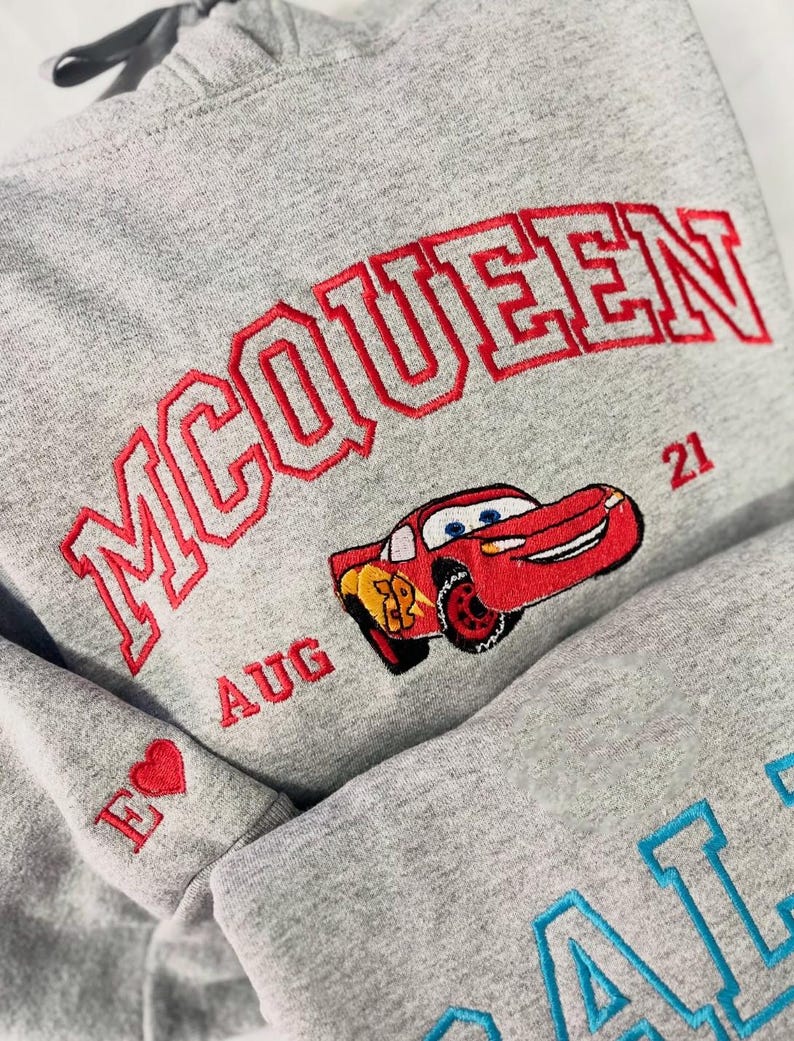 Pode incluir: Um moletom cinza com texto bordado em vermelho lendo "McQUEEN" acima de um carro de desenho animado vermelho. A manga apresenta um cora&ccedil;&atilde;o vermelho e a letra "E". A palavra "AUG" e o n&uacute;mero "21" tamb&eacute;m s&atilde;o bordados no moletom.