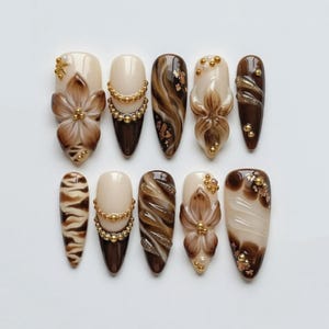 May include: A set of ten almond-shaped press-on nails featuring a color palette of cream, mocha, and dark brown. The designs include hand-painted floral motifs, gold bead accents, swirling marble patterns, tiger stripes, and gold foil flakes.
