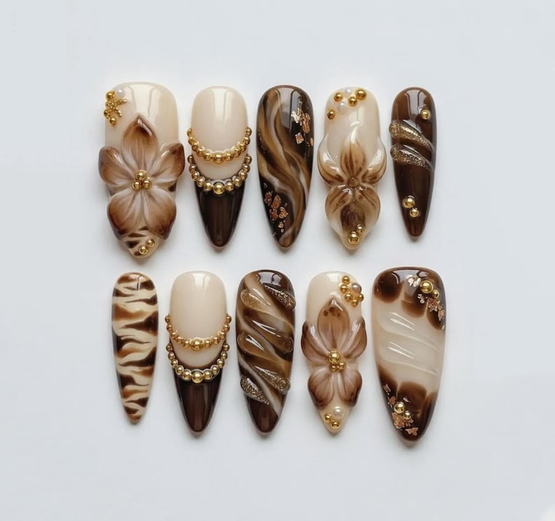 May include: A set of ten almond-shaped press-on nails featuring a color palette of cream, mocha, and dark brown. The designs include hand-painted floral motifs, gold bead accents, swirling marble patterns, tiger stripes, and gold foil flakes.