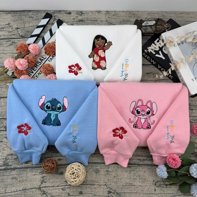 May include: Four folded sweatshirts in white, blue, and pink, each embroidered with a different character from a cartoon. The white sweatshirt features a girl in a red dress, while the blue and pink sweatshirts have cartoon characters. Each sweatshirt has a floral design.