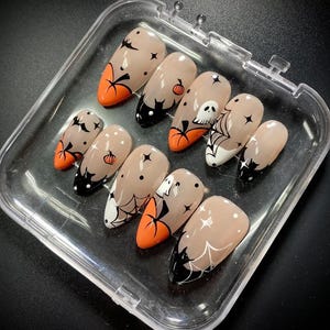 以下が含まれることがあります： ヌードカラーのベースに、黒いコウモリ、オレンジのカボチャ、白い幽霊、黒いクモの巣などのハロウィーンをテーマにしたデザインが施された、アーモンド型のプレスオンネイル10本セット。小さな黒い点や四芒星の形がアクセントになっています。