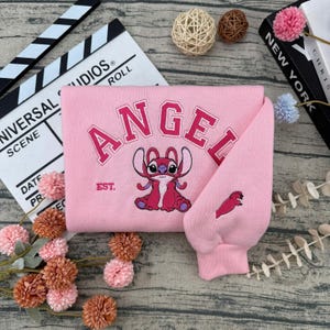 May include: Pink sweatshirt with the word "ANGEL" embroidered in a collegiate font, along with a cartoon character illustration. The sleeve features a small embroidered design. The sweatshirt has the text "EST." below the word "ANGEL".