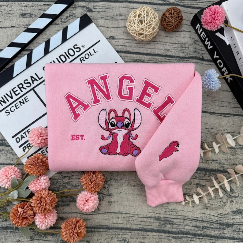 May include: Pink sweatshirt with the word "ANGEL" embroidered in a collegiate font, along with a cartoon character illustration. The sleeve features a small embroidered design. The sweatshirt has the text "EST." below the word "ANGEL".