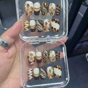May include: A set of press-on nails in a clear plastic case, featuring a color palette of white, dark brown, and gold. The designs include hand-painted brown flowers, gold bead accents, swirling marble patterns, and gold foil flakes on various nails.