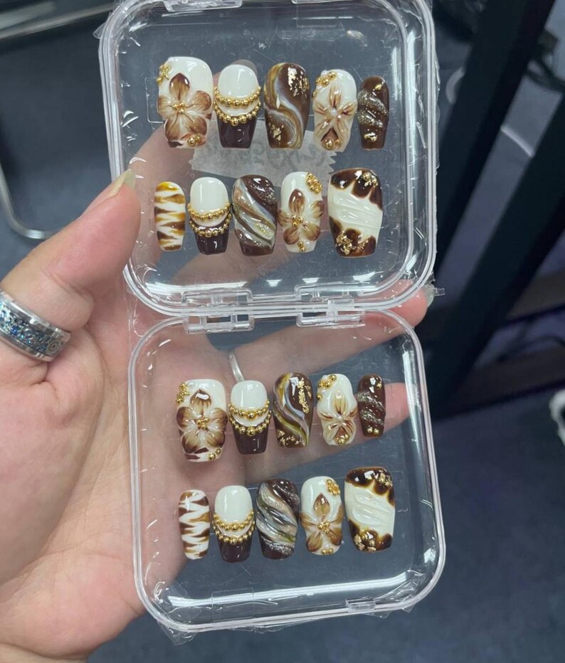May include: A set of press-on nails in a clear plastic case, featuring a color palette of white, dark brown, and gold. The designs include hand-painted brown flowers, gold bead accents, swirling marble patterns, and gold foil flakes on various nails.