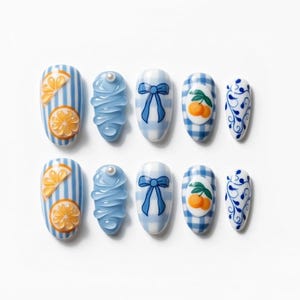 May include: A set of ten artificial nails with various designs. The designs include orange slices, blue bows, gingham patterns with oranges, and swirling blue patterns. The nails are oval-shaped and set against a white background.
