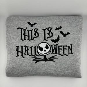 K&ouml;nnte beinhalten: Ein graues Sweatshirt mit dem schwarzen Schriftzug "THIS IS HALLOWEEN". Das Design umfasst Flederm&auml;use und eine von Jack Skellington inspirierte Figur. Das Sweatshirt ist gefaltet.