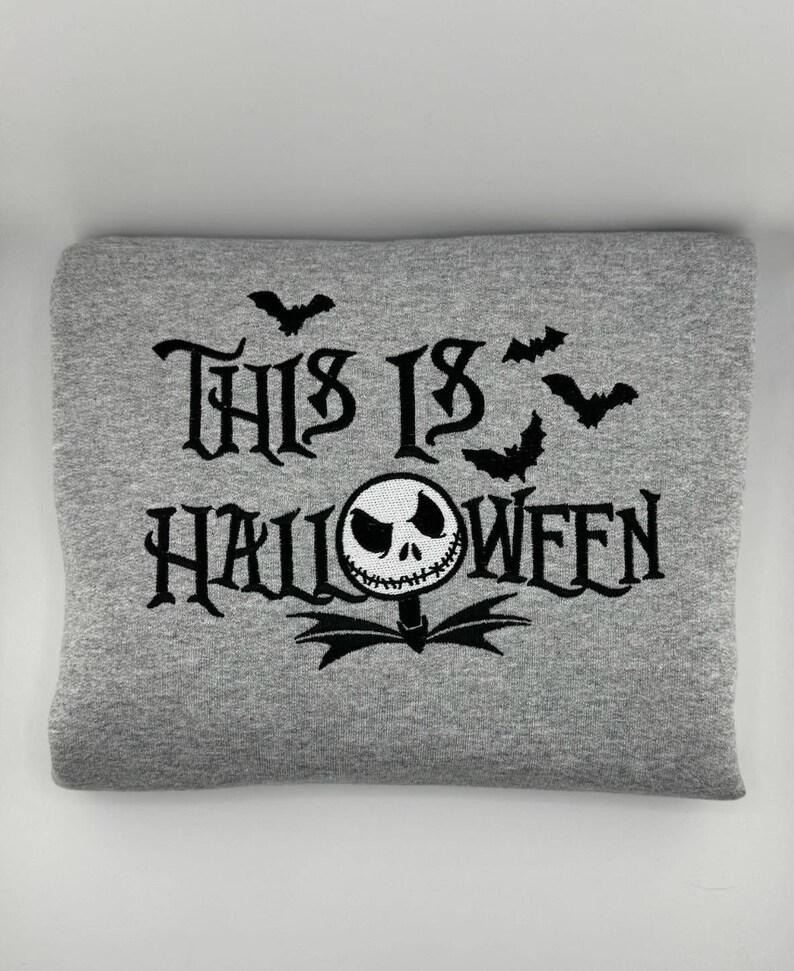 K&ouml;nnte beinhalten: Ein graues Sweatshirt mit dem schwarzen Schriftzug "THIS IS HALLOWEEN". Das Design umfasst Flederm&auml;use und eine von Jack Skellington inspirierte Figur. Das Sweatshirt ist gefaltet.