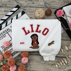 May include: White sweatshirt featuring the name "LILO" in red block letters above an embroidered image of a cartoon character. The sweatshirt also has the word "EST." and a brown embroidered design on the sleeve.