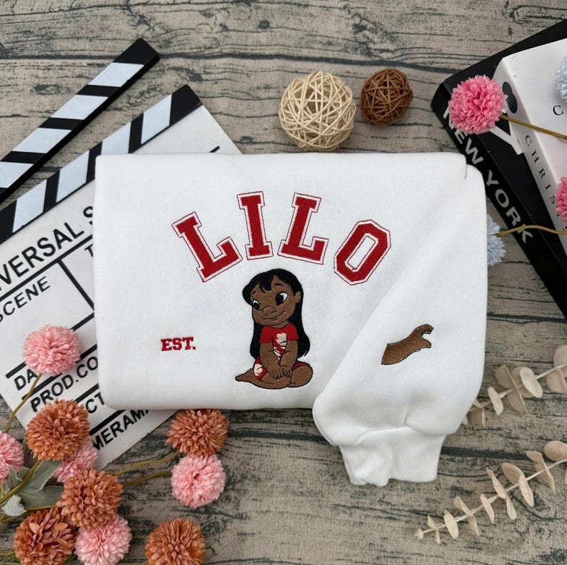 May include: White sweatshirt featuring the name "LILO" in red block letters above an embroidered image of a cartoon character. The sweatshirt also has the word "EST." and a brown embroidered design on the sleeve.