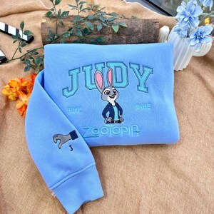 May include: Light blue sweatshirt with the name "JUDY" embroidered in a collegiate font, along with a cartoon image of a rabbit and the text "ZOOTOPIA". The sleeve features a hand graphic. "EST. 2016" is also embroidered on the sweatshirt.