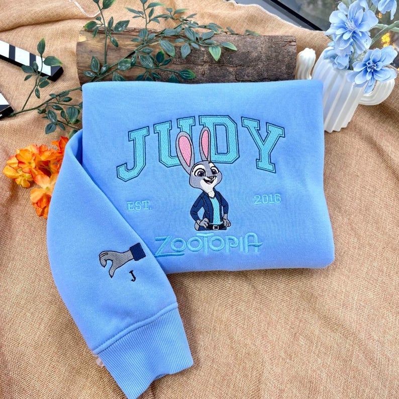 May include: Light blue sweatshirt with the name "JUDY" embroidered in a collegiate font, along with a cartoon image of a rabbit and the text "ZOOTOPIA". The sleeve features a hand graphic. "EST. 2016" is also embroidered on the sweatshirt.