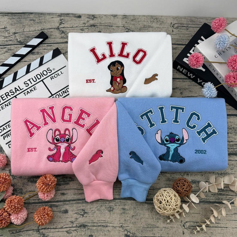 May include: Three sweatshirts in white, pink, and blue, each featuring embroidered cartoon characters and text. The white sweatshirt says "LILO" with a character illustration. The pink sweatshirt says "ANGEL". The blue sweatshirt says "STITCH".