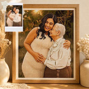 May include: A framed cross-stitch portrait of two people embracing, with a smaller photo in the upper left corner. The portrait features warm tones, with the subjects in light-coloured clothing. The frame is made of light-coloured wood.