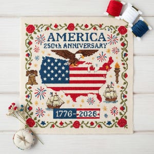 May include: Cross-stitch embroidery featuring the United States flag and map, with an eagle, ships, and the text "AMERICA 250th ANNIVERSARY 1776-2026." The design is framed with floral and starburst motifs, and includes red, white, and blue thread spools.