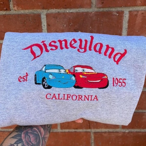 Puede incluir: Una sudadera gris con la palabra "Disneyland" en rojo. Debajo, dos coches de dibujos animados, uno azul y otro rojo. El texto "est 1955 CALIFORNIA" tambi&eacute;n est&aacute; bordado en rojo.