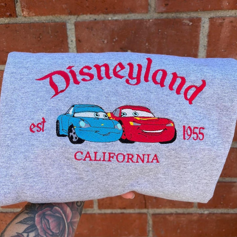 Puede incluir: Una sudadera gris con la palabra "Disneyland" en rojo. Debajo, dos coches de dibujos animados, uno azul y otro rojo. El texto "est 1955 CALIFORNIA" tambi&eacute;n est&aacute; bordado en rojo.
