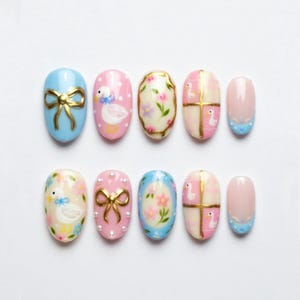 May include: A set of ten oval-shaped false nails. The nails feature various designs, including a gold bow, a duck with a blue bow, floral patterns, and a pink and gold grid pattern. The color palette includes light blue, pink, gold, and white.