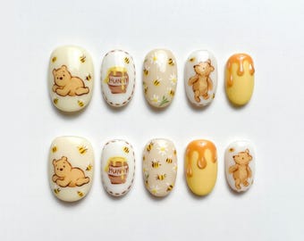 May include: A set of ten press-on nails with a Winnie the Pooh theme. The nails feature designs of Pooh Bear, honey pots, bees, and honey drips in shades of yellow, beige, and brown. The nails are oval-shaped and have a glossy finish.