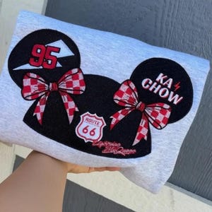 May include: A light gray sweatshirt featuring a large graphic of two black mouse ears with red and white checkered bows. The left ear displays the number 95, while the right ear reads KA CHOW. A Route 66 shield emblem sits between the ears.