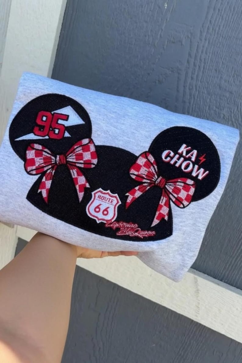 May include: A light gray sweatshirt featuring a large graphic of two black mouse ears with red and white checkered bows. The left ear displays the number 95, while the right ear reads KA CHOW. A Route 66 shield emblem sits between the ears.