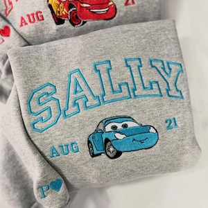 Pode incluir: Duas camisolas cinzentas com desenhos bordados. Uma apresenta "MCQUEEN" em vermelho, um carro vermelho, "AUG 21" e um cora&ccedil;&atilde;o. A outra tem "SALLY" em azul, um carro azul e "AUG 21".