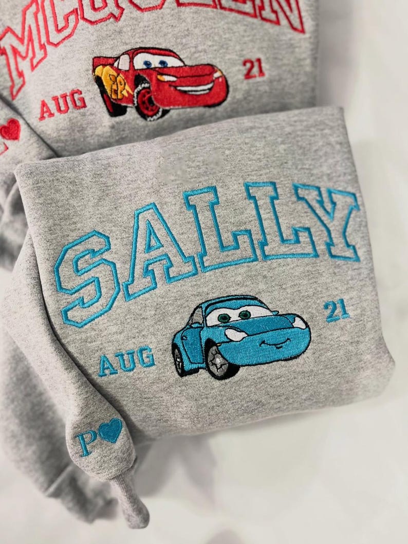 Pode incluir: Duas camisolas cinzentas com desenhos bordados. Uma apresenta "MCQUEEN" em vermelho, um carro vermelho, "AUG 21" e um cora&ccedil;&atilde;o. A outra tem "SALLY" em azul, um carro azul e "AUG 21".