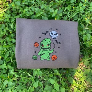 K&ouml;nnte beinhalten: Ein graues Sweatshirt mit einem gestickten Design eines gr&uuml;nen Charakters, der einen Jack Skellington-Ballon h&auml;lt. Das Design umfasst orangefarbene W&uuml;rfel und schwarze Flederm&auml;use. Das Sweatshirt ist auf gr&uuml;nem Gras gefaltet.