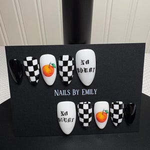 May include: A set of artificial nails with various designs. The nails feature a black and white checkerboard pattern, a realistic orange fruit, and the text "No Doubt". The nails are displayed on a black card with the text "NAILS BY EMILY".