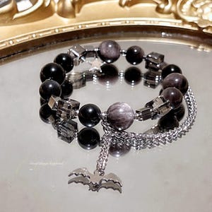 May include: A beaded bracelet featuring alternating black and dark grey spherical beads, translucent dark grey cube beads, and a small silver-toned star charm. A silver-toned chain hangs from the bracelet with a bat-shaped charm. Text reads: Good things happend.