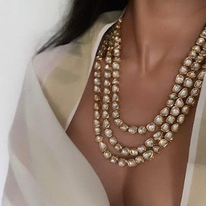 May include: A layered gold necklace featuring multiple strands of large, irregular, white gemstones. The necklace is worn against a cream-coloured garment. The gemstones are set in gold bezels.