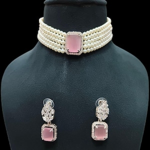 Könnte beinhalten: Eine Perlen-Choker-Halskette mit einem rechteckigen rosa Edelstein in der Mitte und passenden Ohrringen. Die Halskette hat mehrere Stränge aus kleinen, weißen Perlen. Die Ohrringe und der Mittelteil der Halskette sind mit silberfarbenem Metall und kleinen, klaren Steinen umrahmt.