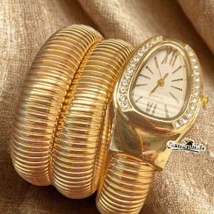 May include: Gold-toned quartz watch with a unique bracelet design. The watch features a white face with Roman numerals and a bezel encrusted with small crystals. The bracelet is made of a series of ribbed, gold-colored segments.