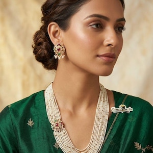 Peut inclure: Un collier de perles à plusieurs rangs avec un pendentif circulaire doré orné de pierres rouges et vertes, assorti à des boucles d'oreilles. L'ensemble est porté avec un vêtement vert foncé. Le texte Gemoraindia est visible sur la droite.