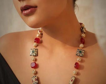 May include: A gold-toned necklace featuring red gemstone beads and ornate square and oval pendants with green and white accents. The necklace is displayed against a neutral background, showcasing its intricate design and craftsmanship.