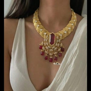 May include: A gold-toned statement necklace featuring a large, rectangular ruby pendant and smaller ruby accents. The necklace has an ornate design with intricate detailing and a bold, eye-catching appearance.