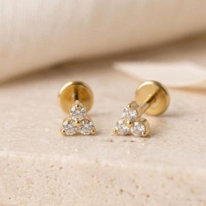 May include: A pair of gold stud earrings, each featuring a cluster of three small, round, clear stones arranged in a triangular pattern. The earrings have a flat, circular back post and a polished gold finish, showcasing a simple and clean design.