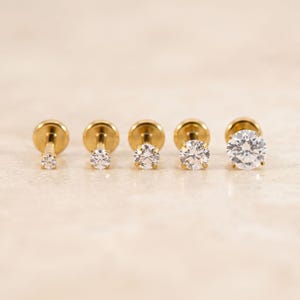 May include: A row of five gold-tone flat back stud earrings arranged by size from smallest to largest. Each earring features a single round, clear, faceted gemstone held in a four-prong setting, attached to a thin gold post with a flat circular base.