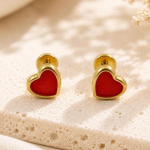 May include: A pair of gold-tone stud earrings featuring a heart-shaped design with a smooth, vibrant red center inlay. The earrings have a polished metallic border and a secure flat-back post mechanism, presenting a clean and simple aesthetic.