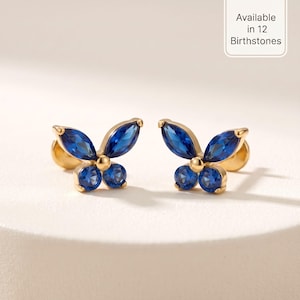 May include: A pair of gold stud earrings shaped like butterflies, each featuring two marquise-cut blue stones for the upper wings and two round-cut blue stones for the lower wings. A small text box in the corner states Available in 12 Birthstones.