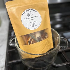 May include: A tan pouch labeled The Cozy Goose co Feathers & Flannels Simmer pot kit containing dried orange slices, cinnamon sticks, rosemary, and star anise, resting inside a clear, ribbed glass pot on a black stovetop burner.