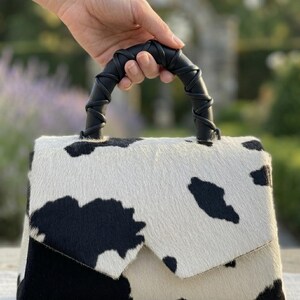 May include: A handbag featuring a black and white cow print pattern with a textured hair-on-hide finish. The bag has a structured flap closure and a black handle wrapped in a decorative leather cord, providing a distinct contrast to the spotted body.