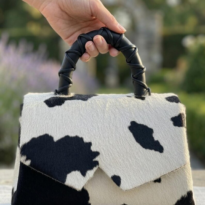 May include: A handbag featuring a black and white cow print pattern with a textured hair-on-hide finish. The bag has a structured flap closure and a black handle wrapped in a decorative leather cord, providing a distinct contrast to the spotted body.