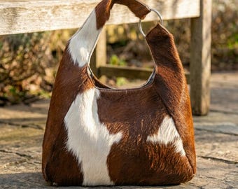 May include: A hobo-style handbag crafted from brown and white cowhide with a natural hair-on texture. The bag features a single integrated handle secured by a circular silver-toned metal ring, showcasing a distinct mottled pattern throughout.