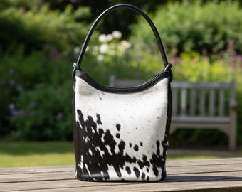 May include: A cowhide bucket bag with a black leather handle and trim. The bag features a white background with black spotted patterns. The bag is sitting on a wooden table in an outdoor setting.