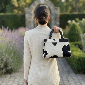 May include: A structured handbag featuring a black and white cow print pattern with a textured finish. The bag has a single top handle with a braided design and a rectangular silhouette. It is paired with a light beige blazer worn over the shoulders.